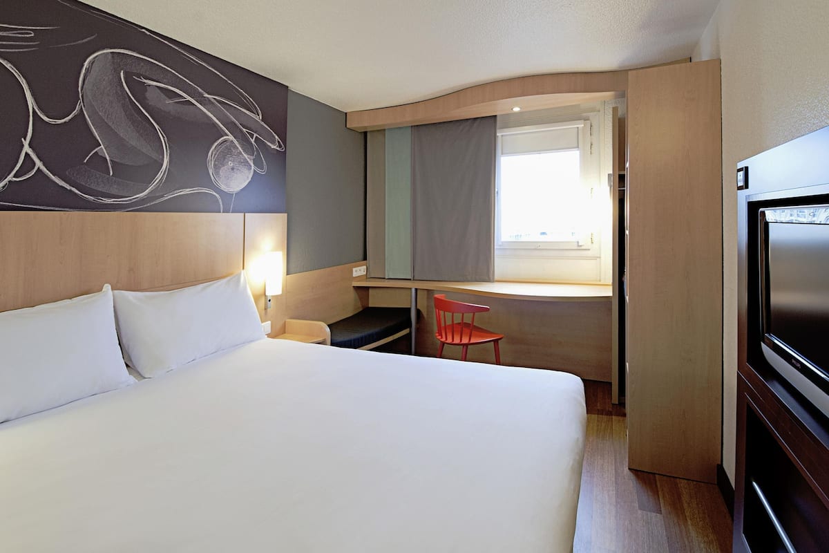 A hotel room with a bed, a desk, a chair, a TV, and a window.