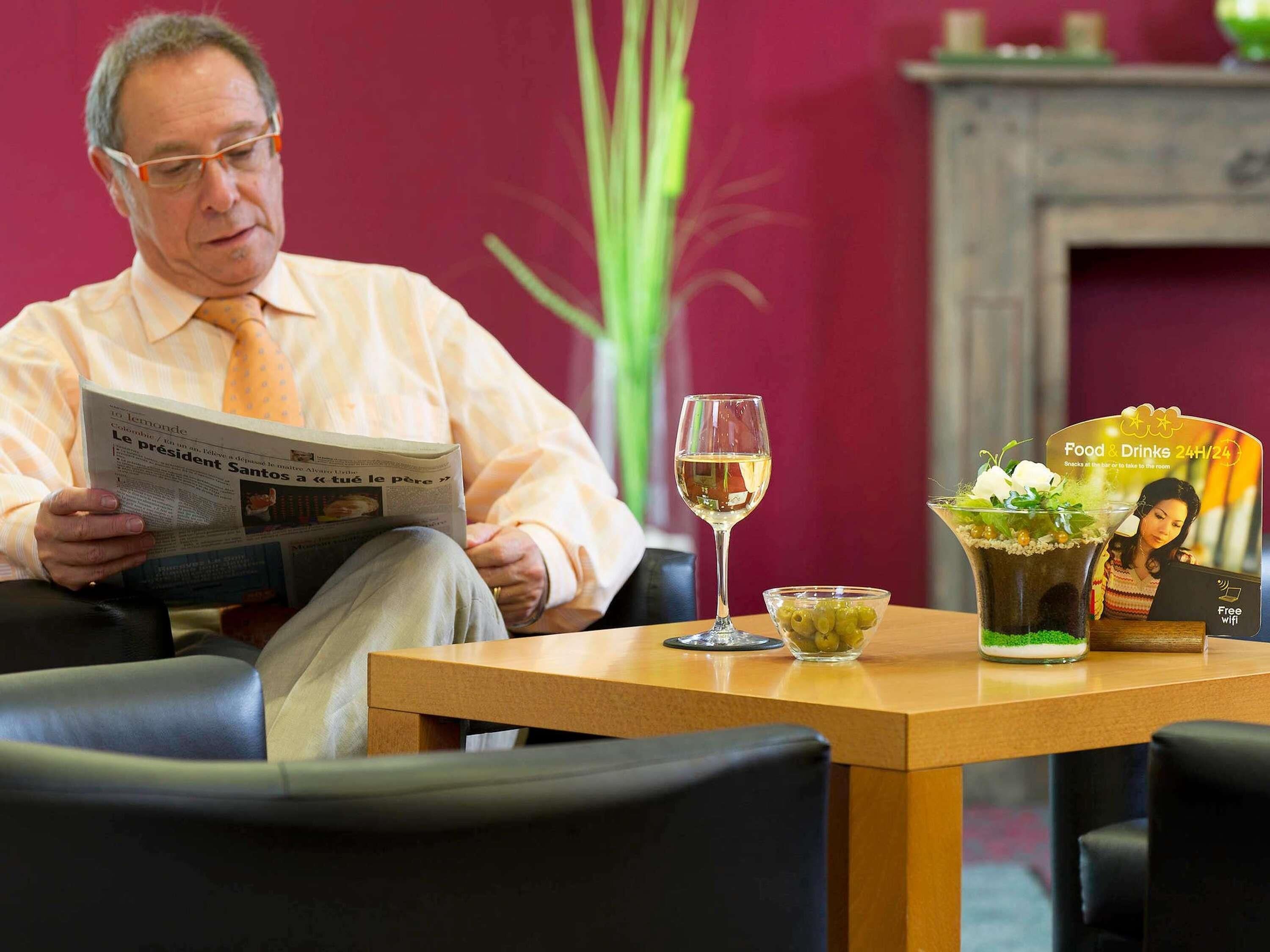 Een man leest een krant in een kamer met een tafel waarop een glas wijn, een schaal olijven en een menukaart staan.
