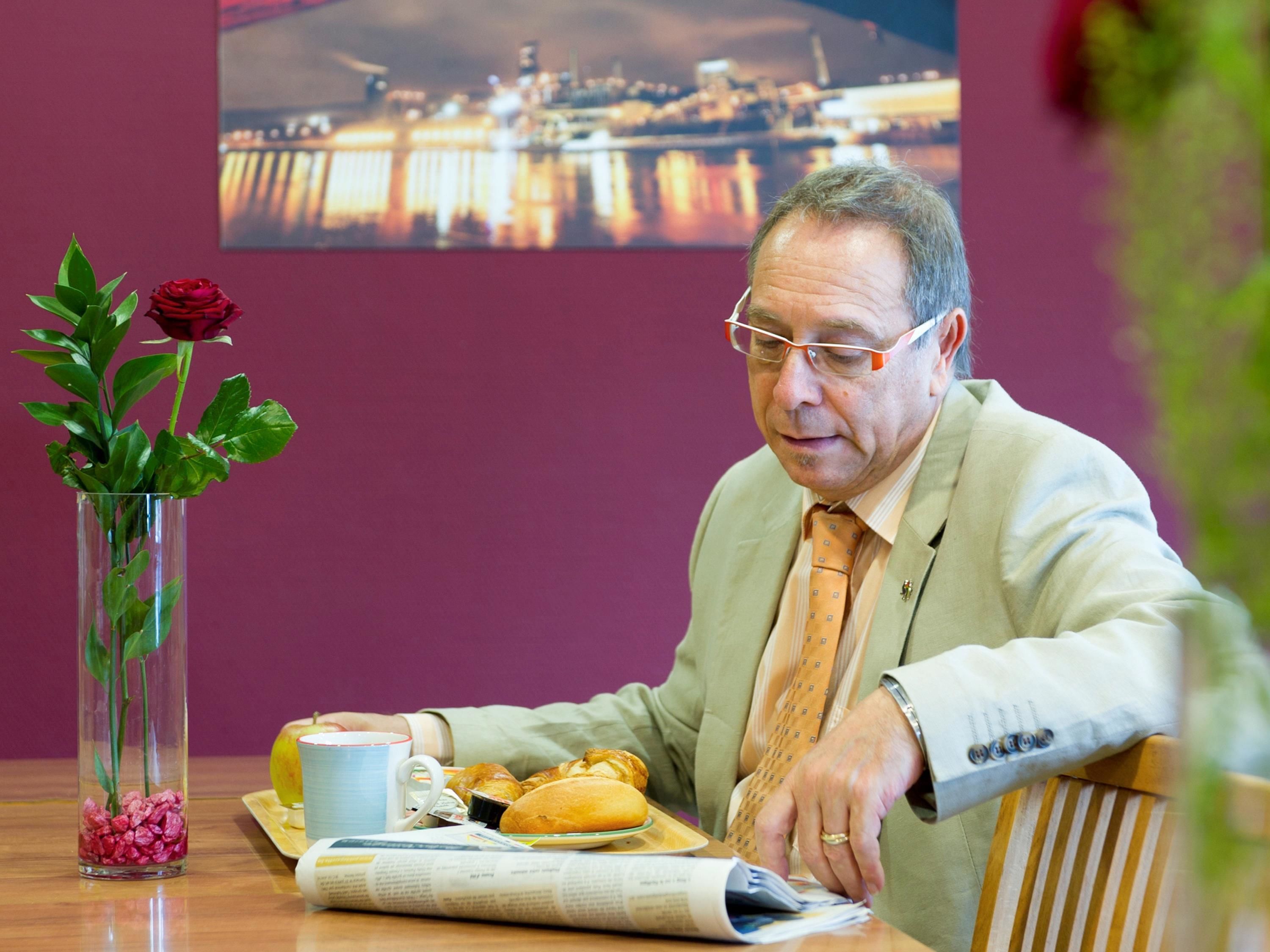 Un hombre sentado a una mesa con comida, un florero con una rosa y un paisaje urbano de fondo.