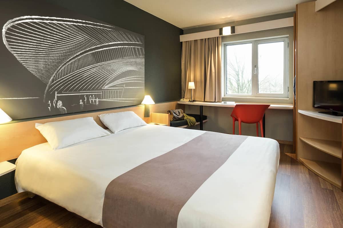 Una habitación de hotel con una cama grande, un escritorio, un televisor y un ventanal con cortinas.