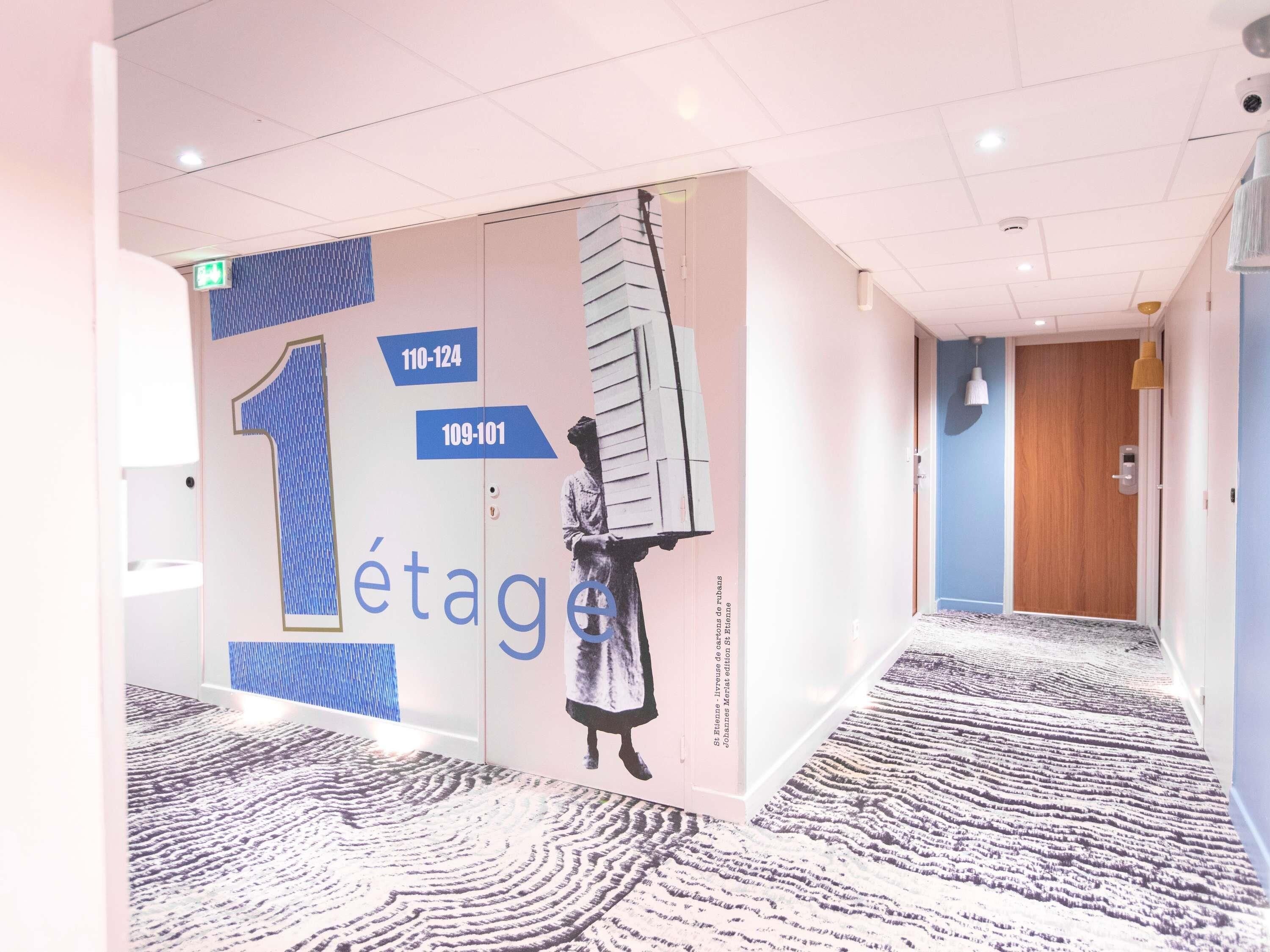 A hotel corridor with a patterned carpet, a wall with a large number '1' and a figure holding a ladder, and a wooden door at the end.