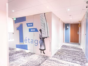 A hotel corridor with a patterned carpet, a wall with a large number '1' and a figure holding a ladder, and a wooden door at the end. - ibis Styles Saint Étienne Gare Châteaucreux (Saint-Etienne)