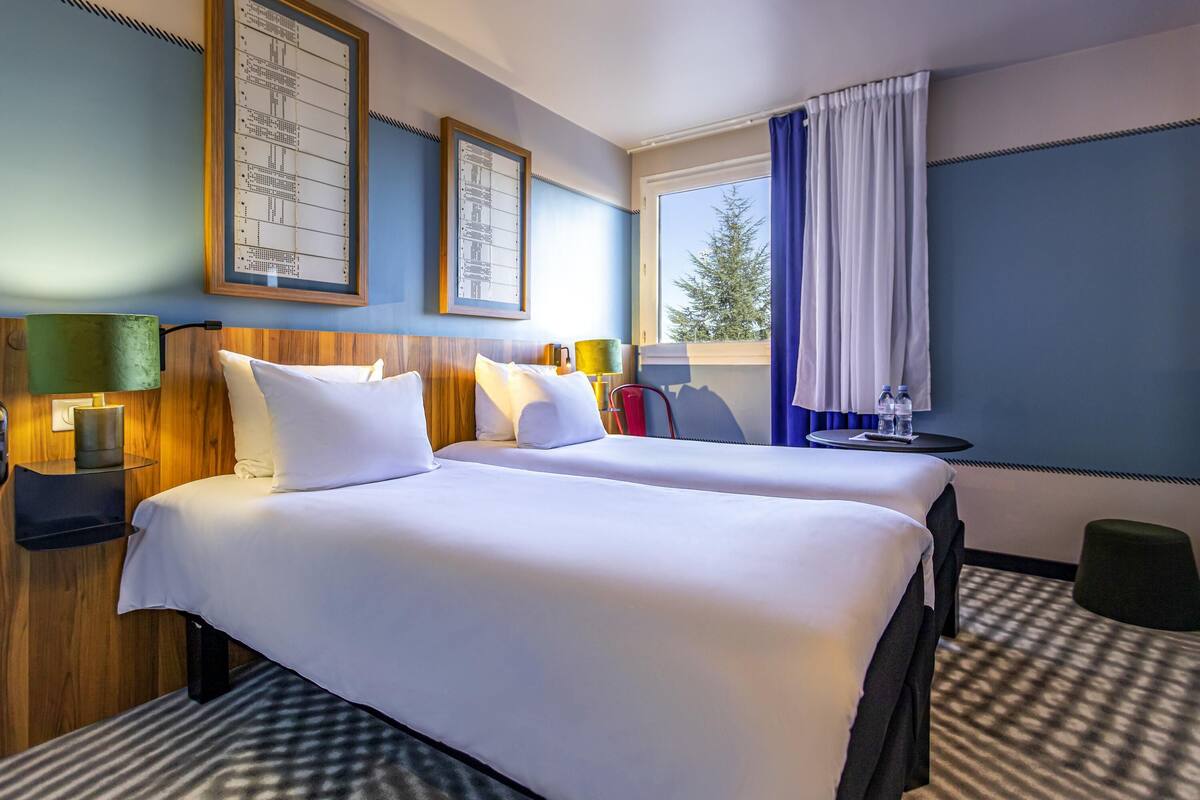 A hotel room with two beds, a window with curtains, a small table with a water bottle, and framed artwork on the wall.