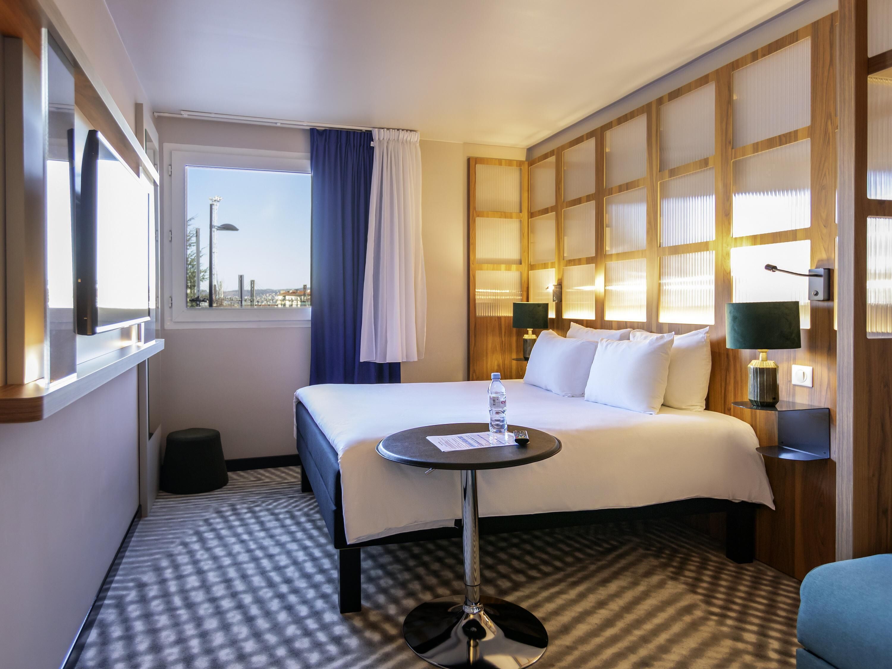 Habitación de hotel con cama, mesita de noche, lámpara y vista a la ciudad a través de una ventana.
