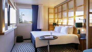 Habitación de hotel con cama, mesita de noche, lámpara y vista a la ciudad a través de una ventana.