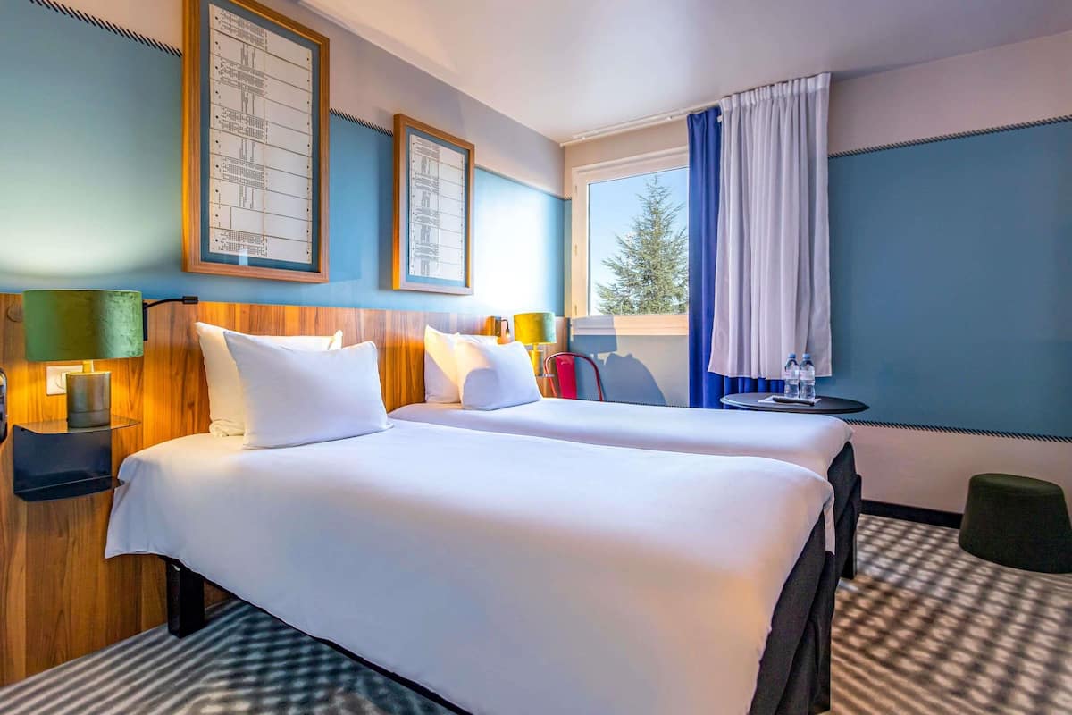 Une chambre d’hôtel avec deux lits, une fenêtre avec des rideaux, une petite table avec des bouteilles d’eau et des œuvres d’art encadrées accrochées au mur.