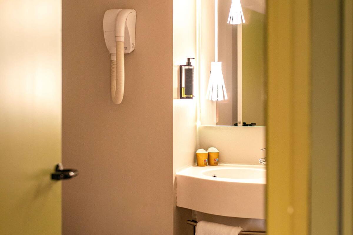 Ett badrum med ett vitt handfat, en spegel, en handduk som hänger på väggen och en hårtork som är monterad på väggen.