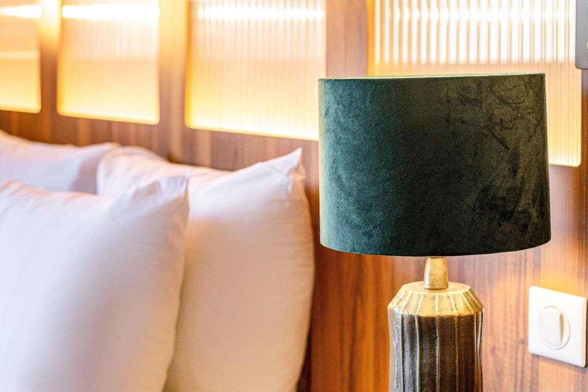 Ein Bett mit weißen Kissen und einer Lampe mit strukturierter Schirm auf einem Nachttisch aus Holz.