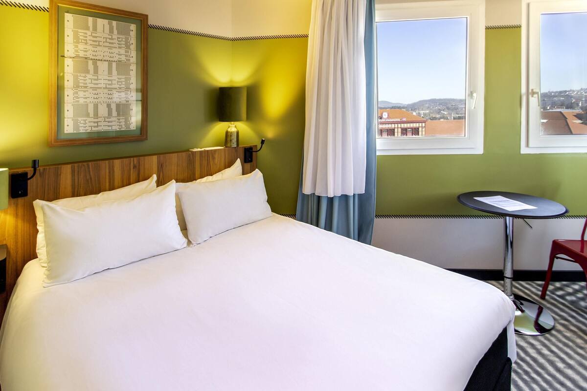 Ett hotellrum med en säng, ett litet bord, en stol, en lampa och ett fönster med utsikt över byggnader.