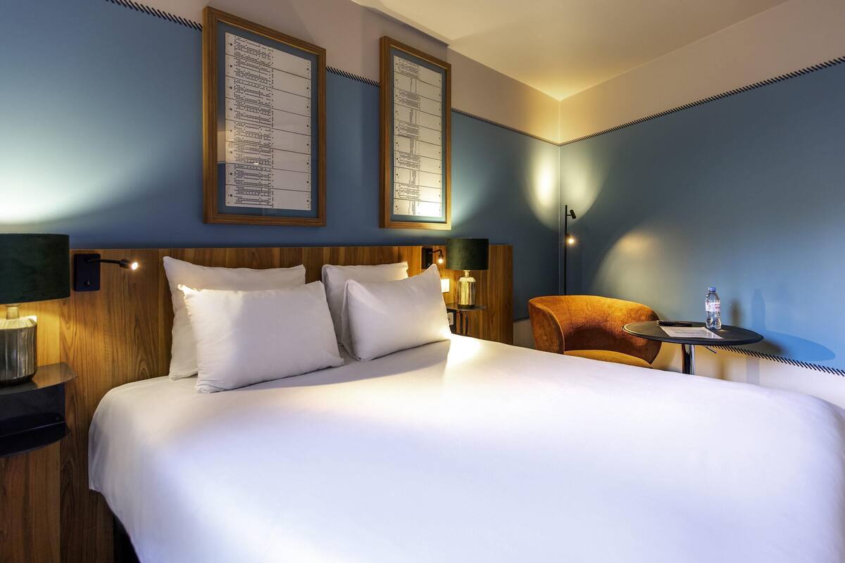 A hotel room with a large bed, a wooden headboard, a nightstand with a lamp, a small table with a bottle, and a chair.