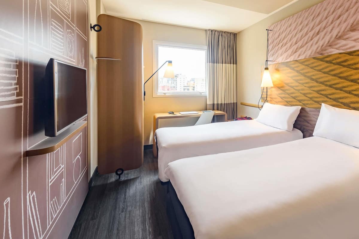 A hotel room with two beds, a desk, a TV, and a window with curtains.