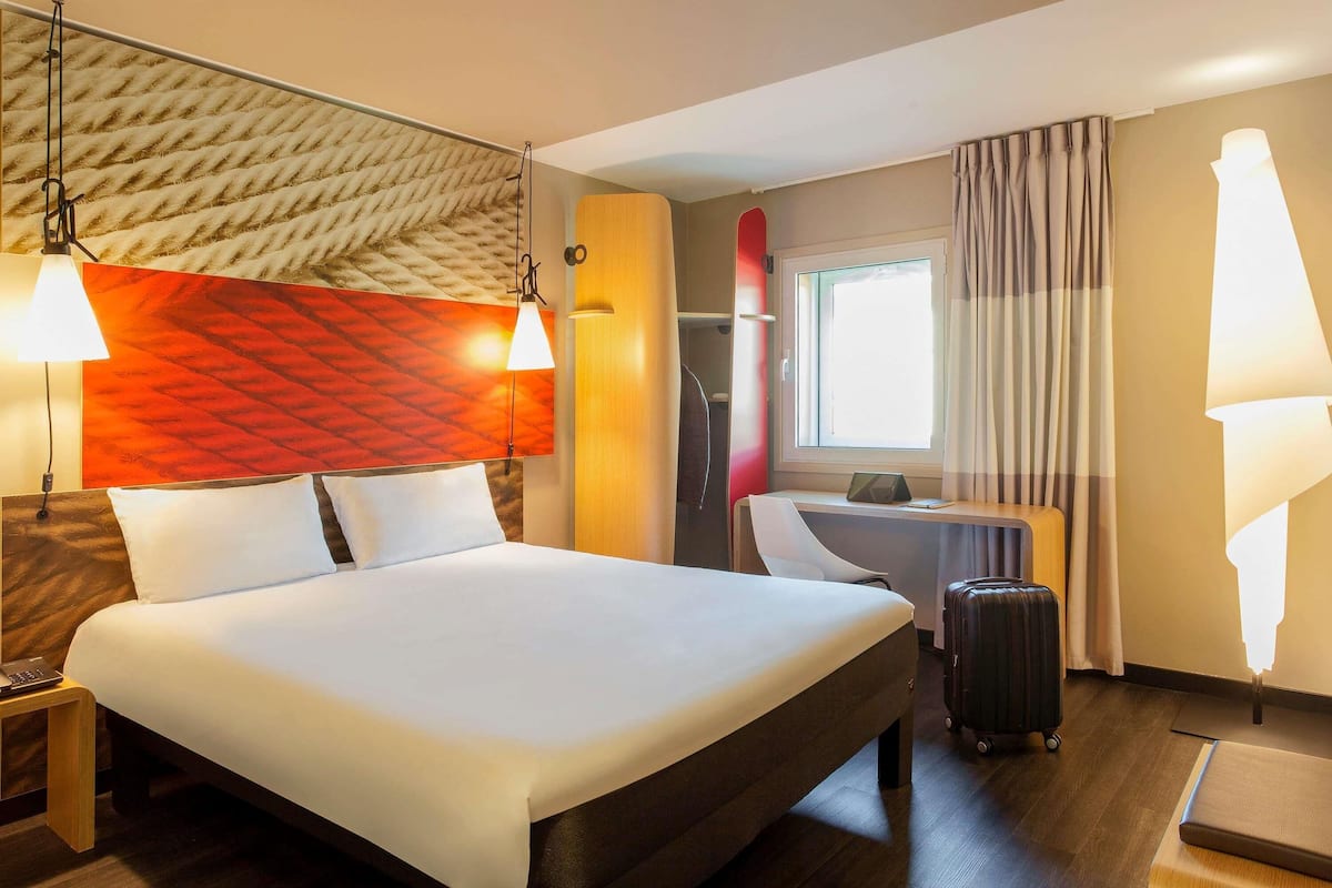 A hotel room with a bed, desk, chair, suitcase, and a window with curtains.