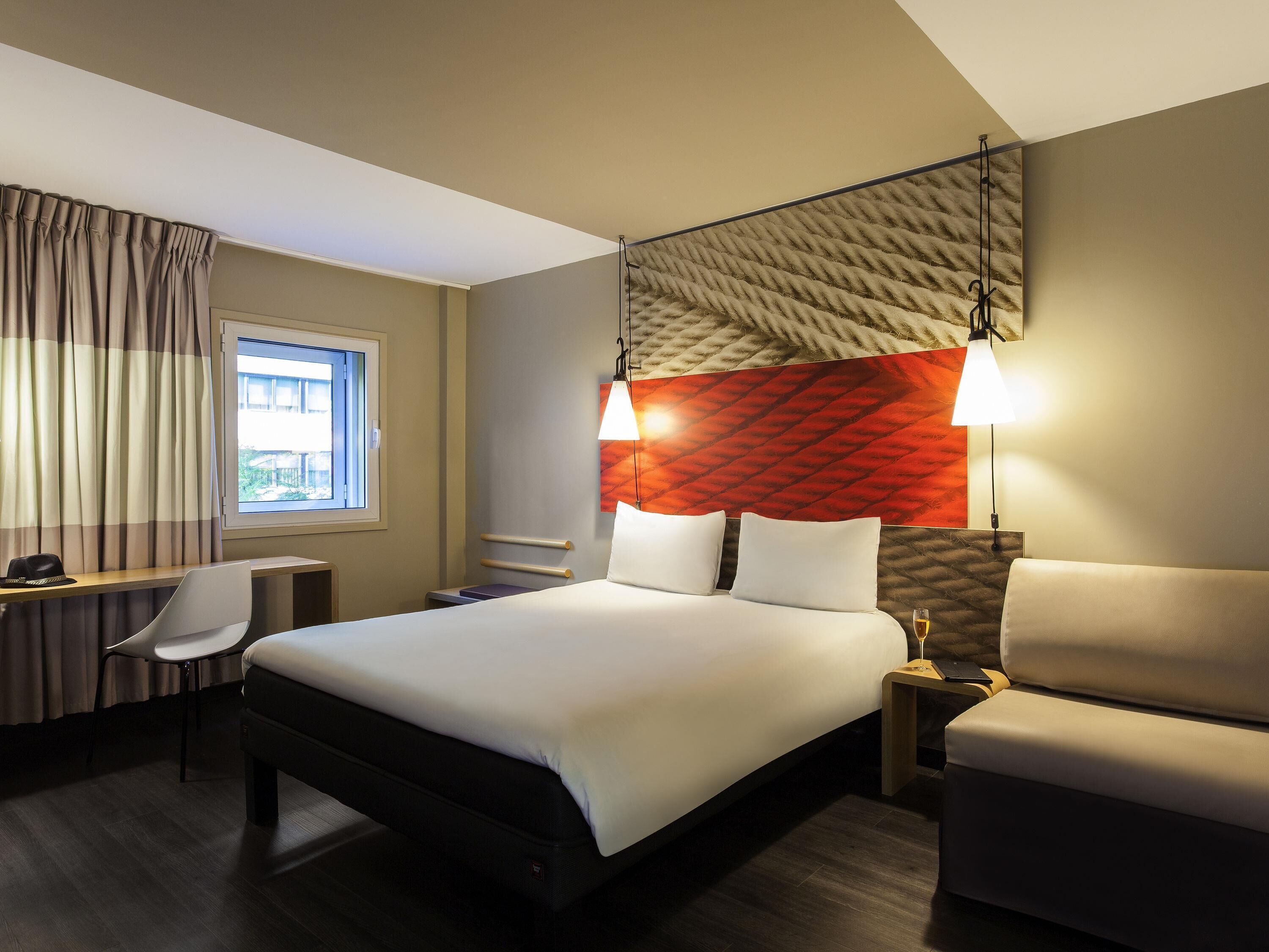 A hotel room with a bed, two bedside tables, a desk with a chair, a window with curtains, and a wall-mounted lamp.
