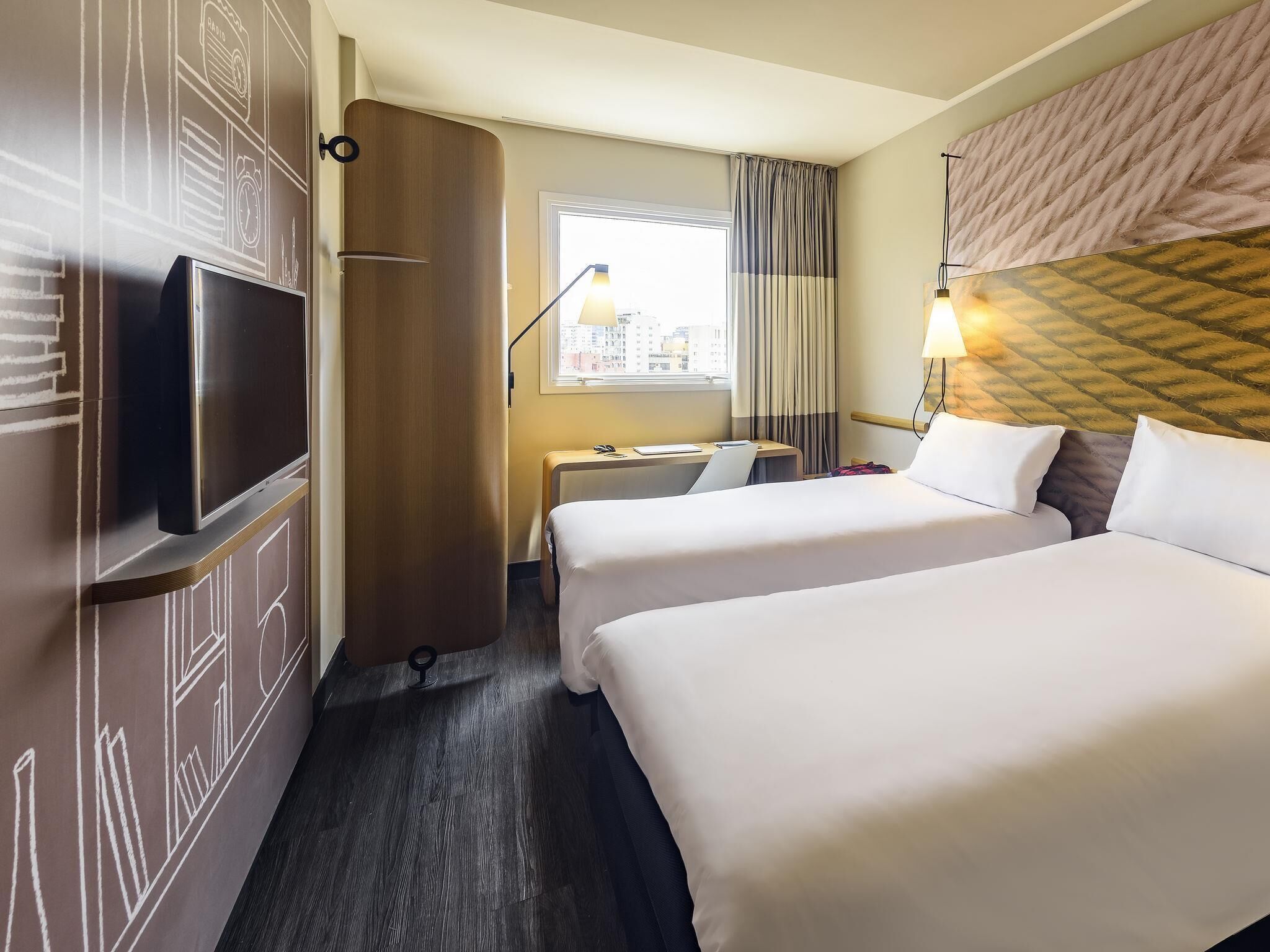 A hotel room with two beds, a desk, a TV, and a window with curtains.