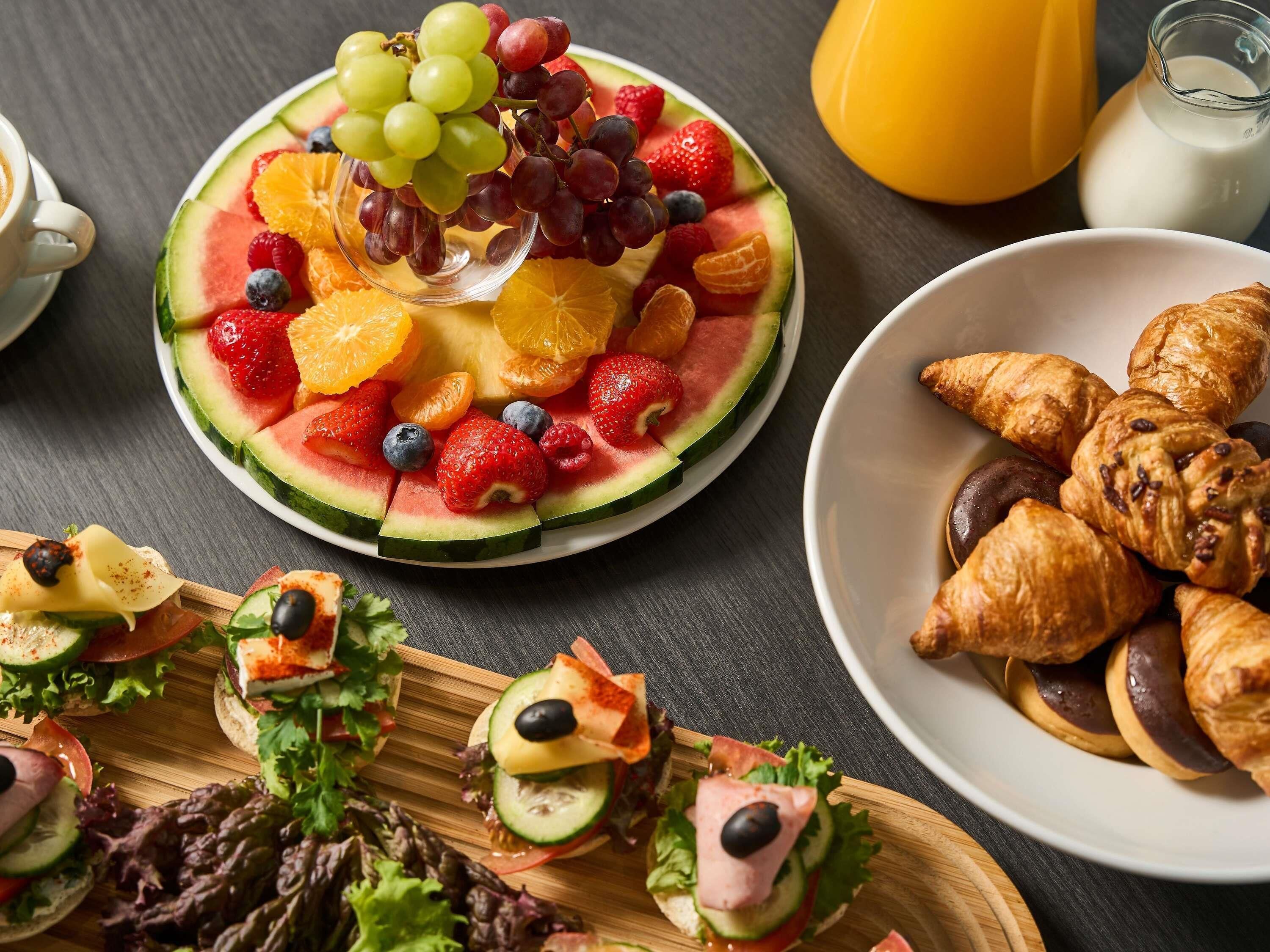 Une table dressée pour le petit-déjeuner, avec une tranche de pastèque garnie de fruits, un bol de croissants et une assiette de sandwichs.