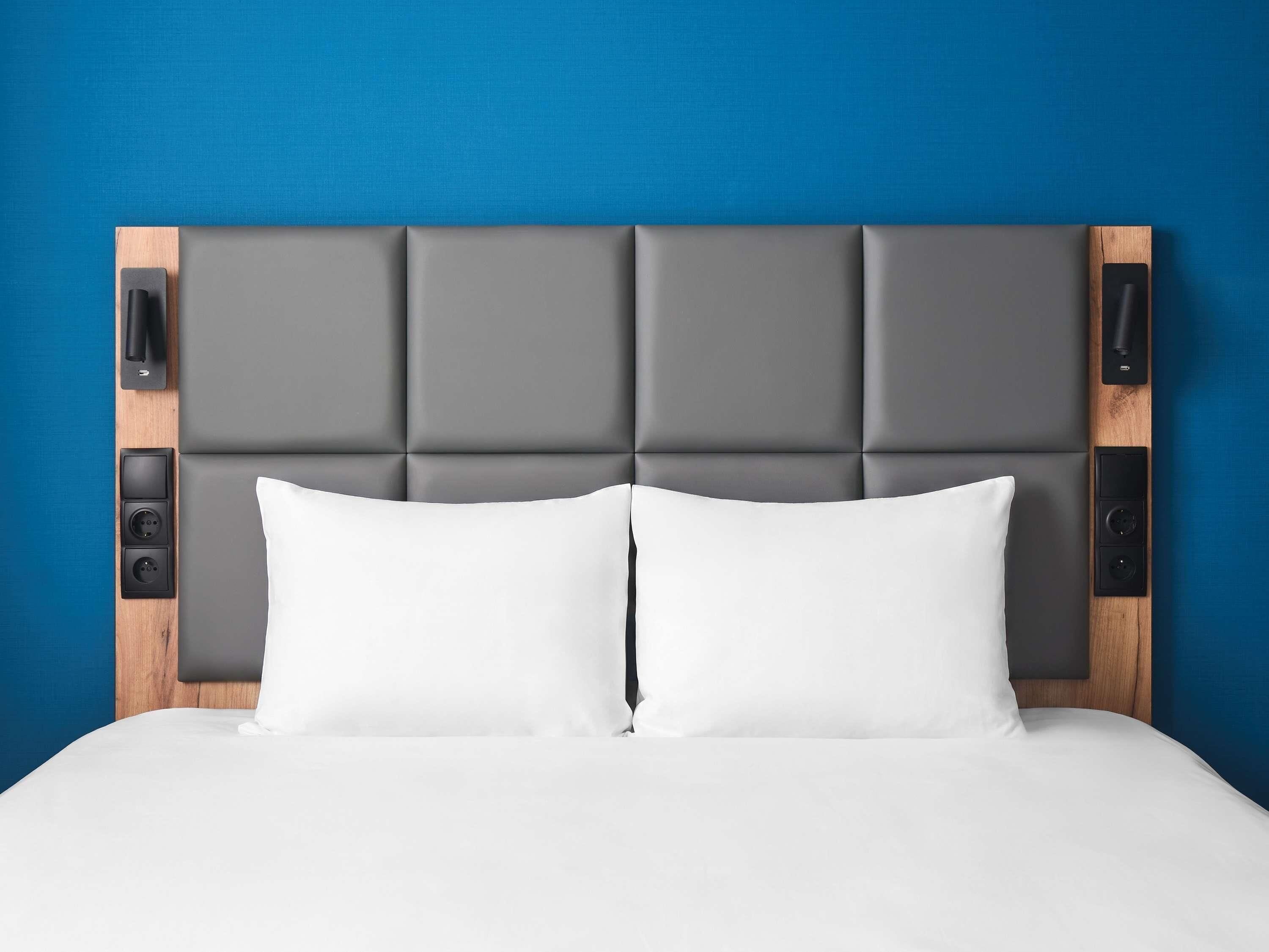 A bed with a grey upholstered headboard, white pillows, and a wooden panel with a control panel on the left side.