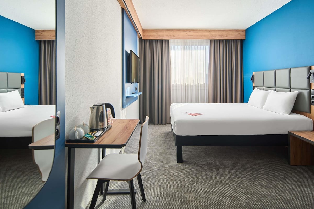 Habitación de hotel con dos camas, un escritorio con silla, un televisor montado en la pared y una ventana con cortinas.