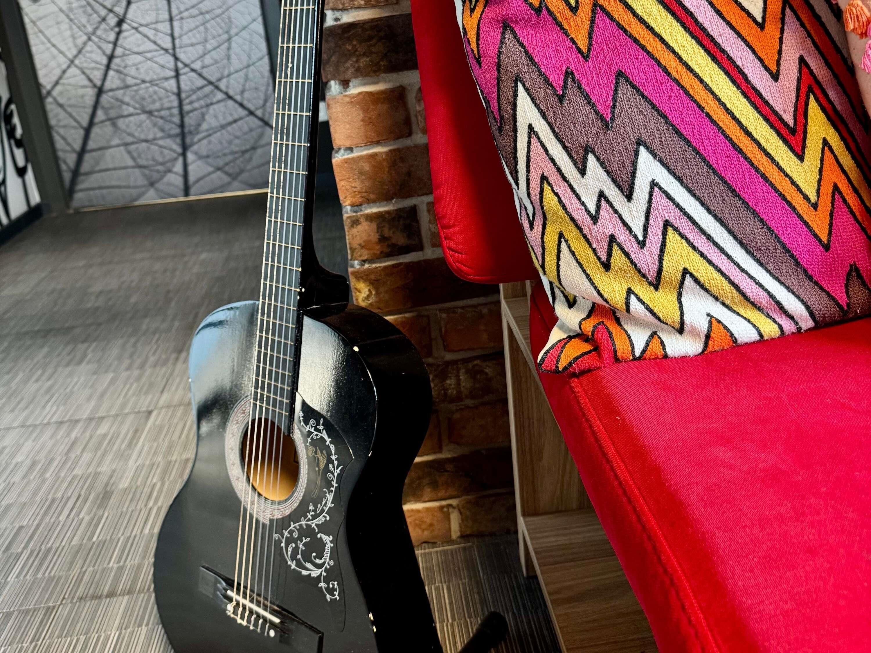 A black guitar leaning against a brick wall with a colorful zigzag patterned cushion on a red chair.