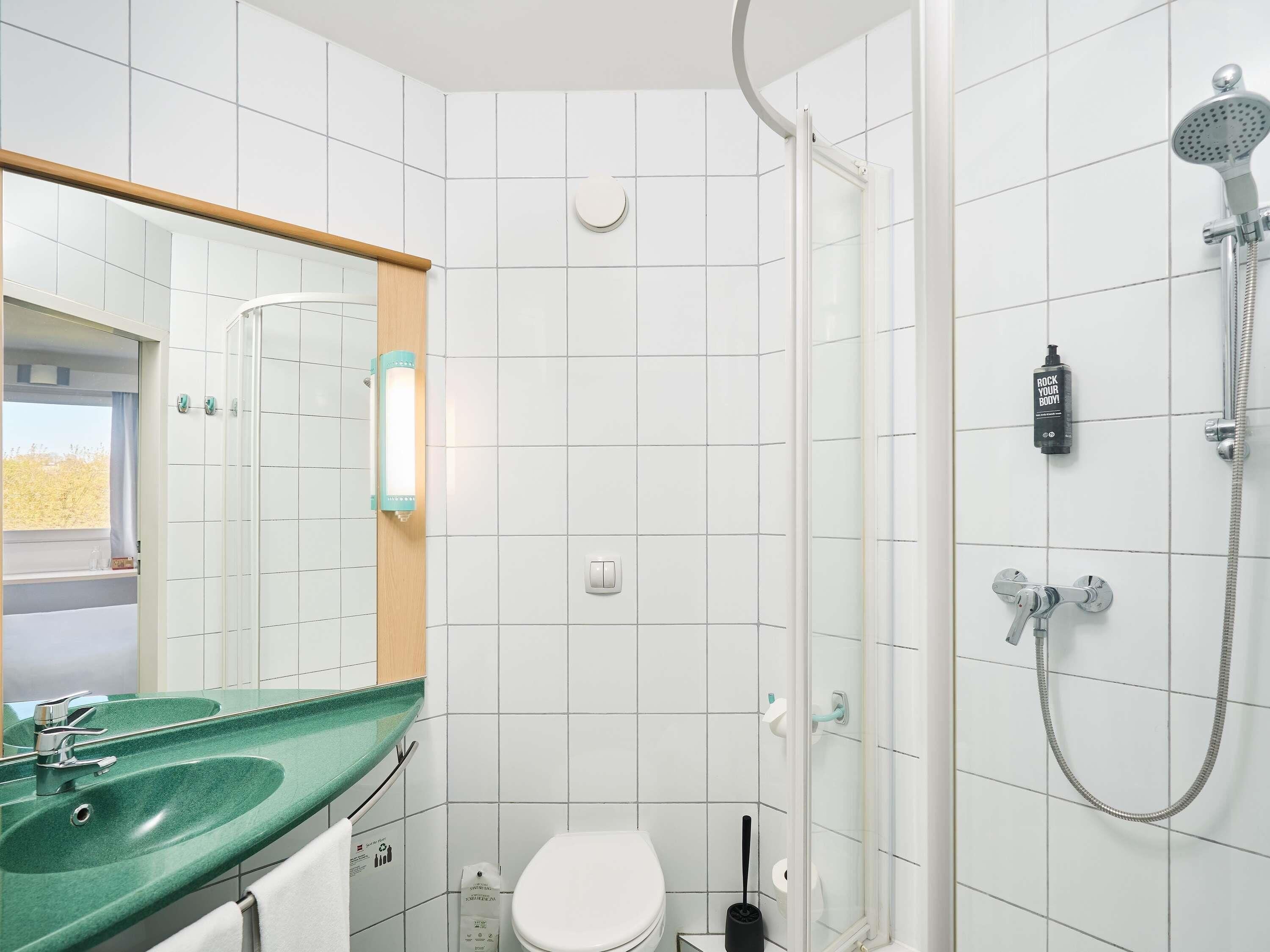 Un baño con un lavabo verde, un inodoro blanco, una ducha con cabezal de ducha manual y un espejo con marco de madera.