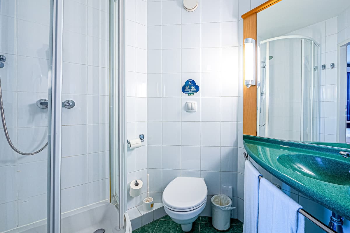 Baño con ducha de vidrio, inodoro, cubo de basura y lavamanos verde con espejo arriba.