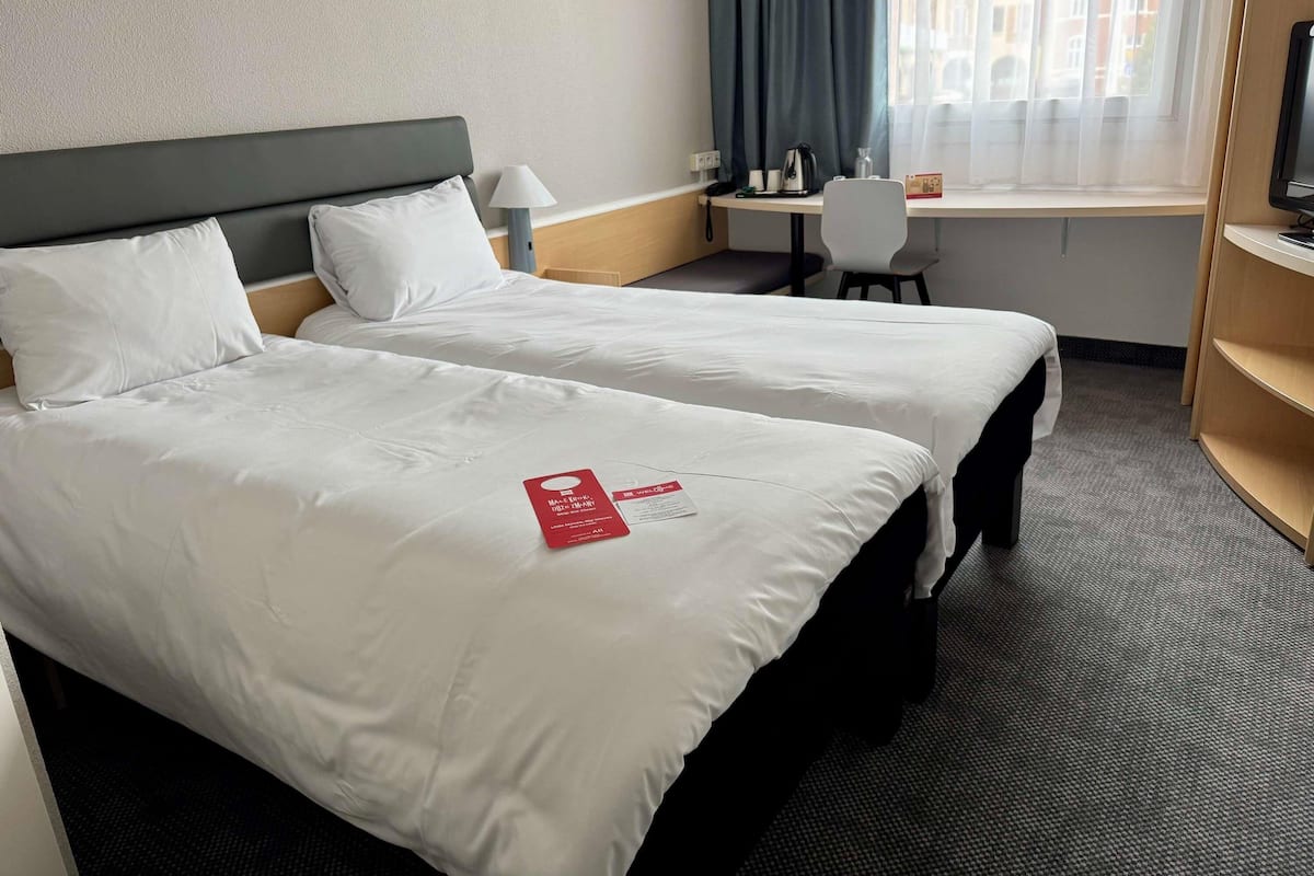 Habitación de hotel con dos camas, un escritorio, una silla, una lámpara y una ventana con cortinas.