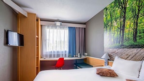 A hotel room with a bed, a desk, a red chair, a television, and a forest view.
