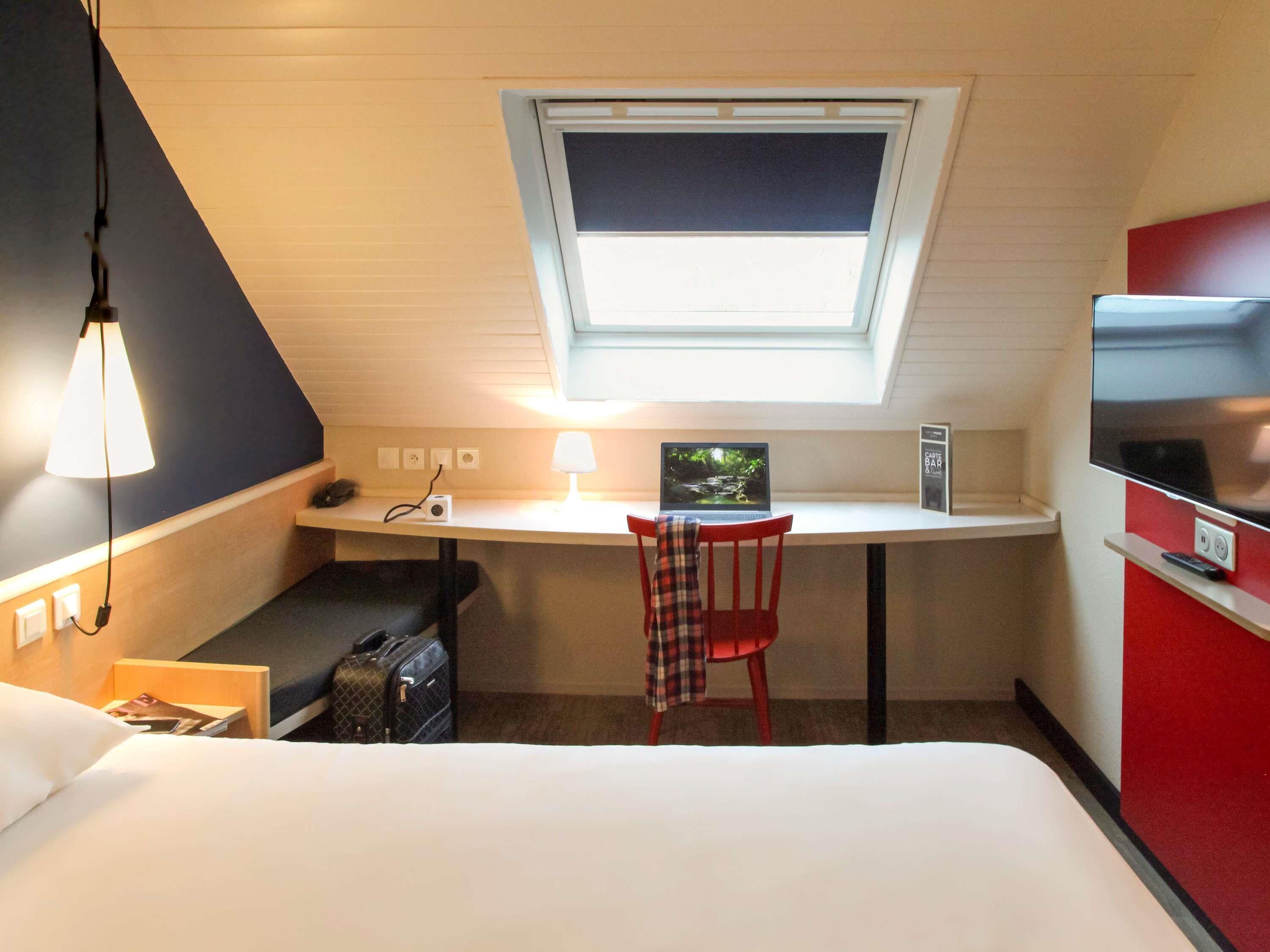 A hotel room with a desk, a laptop, a chair, a bed, a suitcase, and a TV.