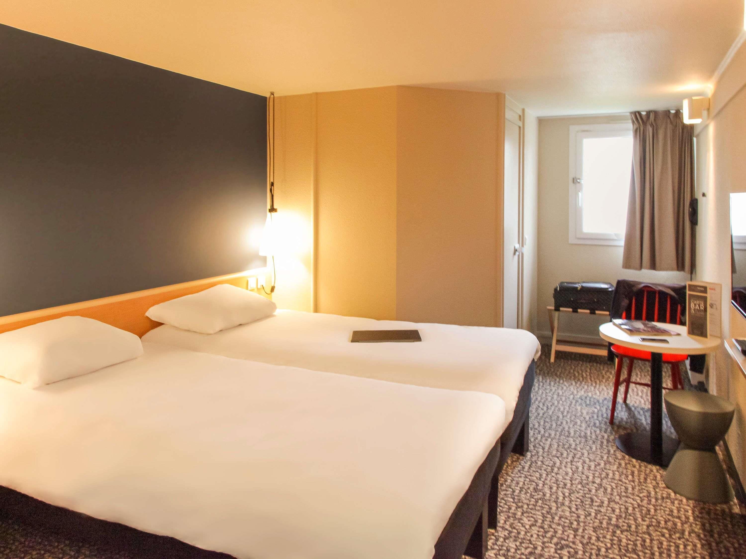 A hotel room with a large bed, a desk, a chair, a small table, and a window with curtains.