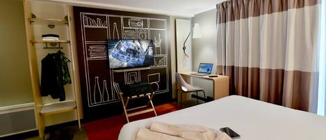 Una habitación de hotel con cama, escritorio con computadora, televisor montado en la pared y armario.