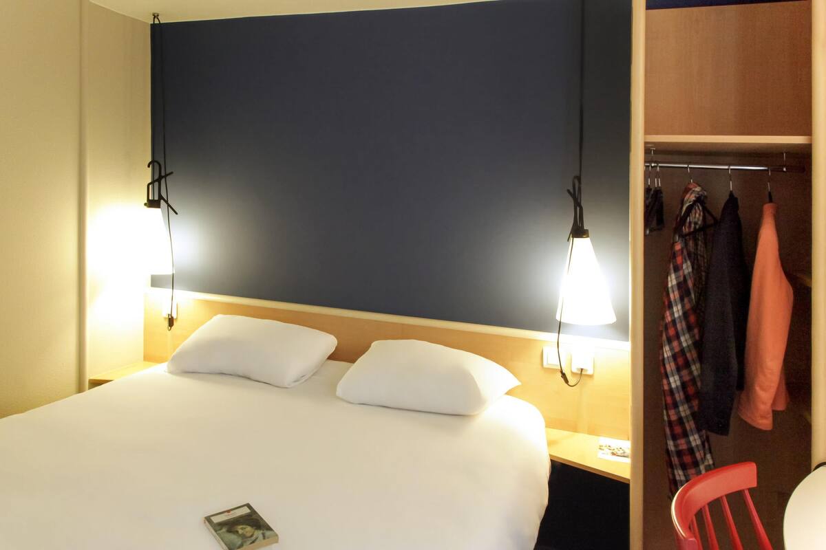 A hotel room with a bed, bedside lamps, a nightstand, a red chair, and a coat rack with clothes.
