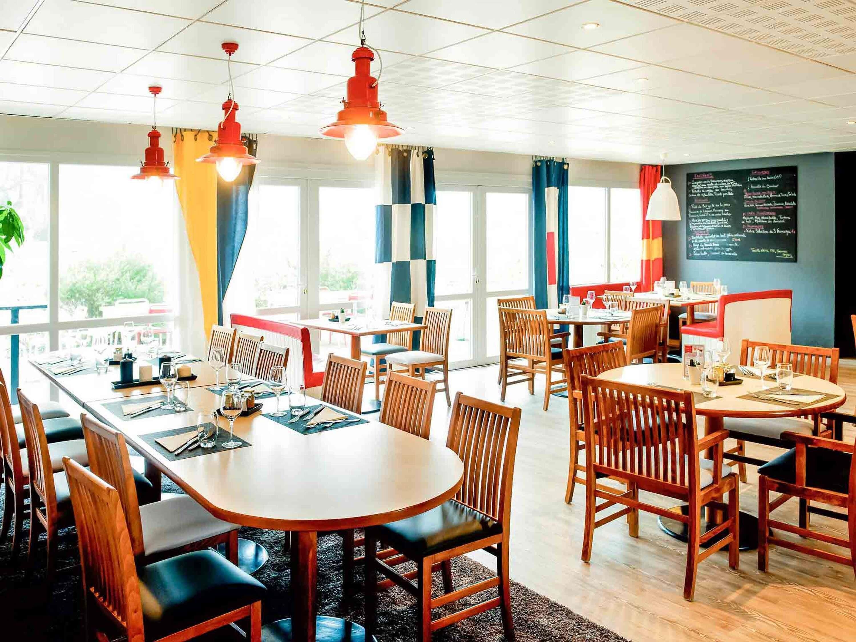 Un restaurant doté de nombreuses tables et chaises, de grandes fenêtres et de luminaires suspendus.