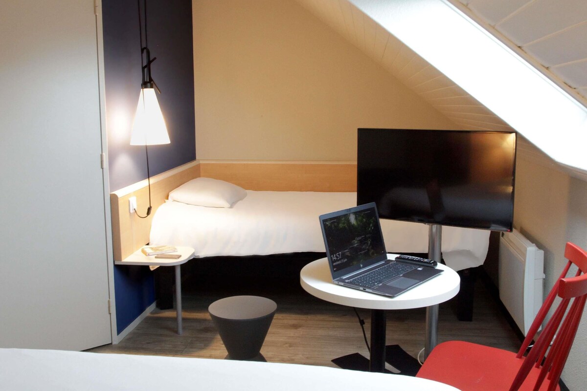 A compact hotel room with a bed, a desk with a laptop, a chair, and a small table.