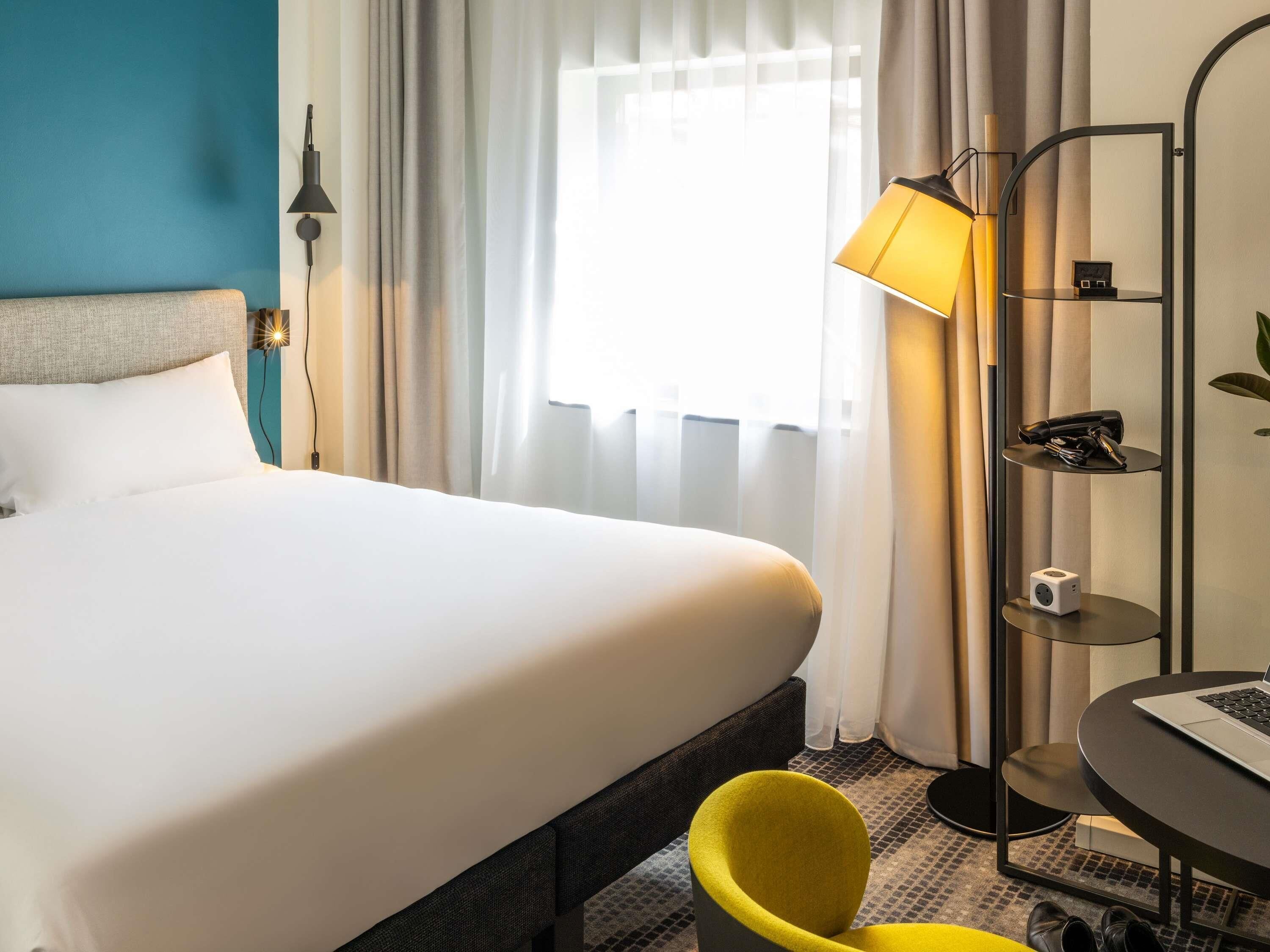 A hotel room with a bed, a desk with a laptop, a chair, a lamp, and a shelf with items.