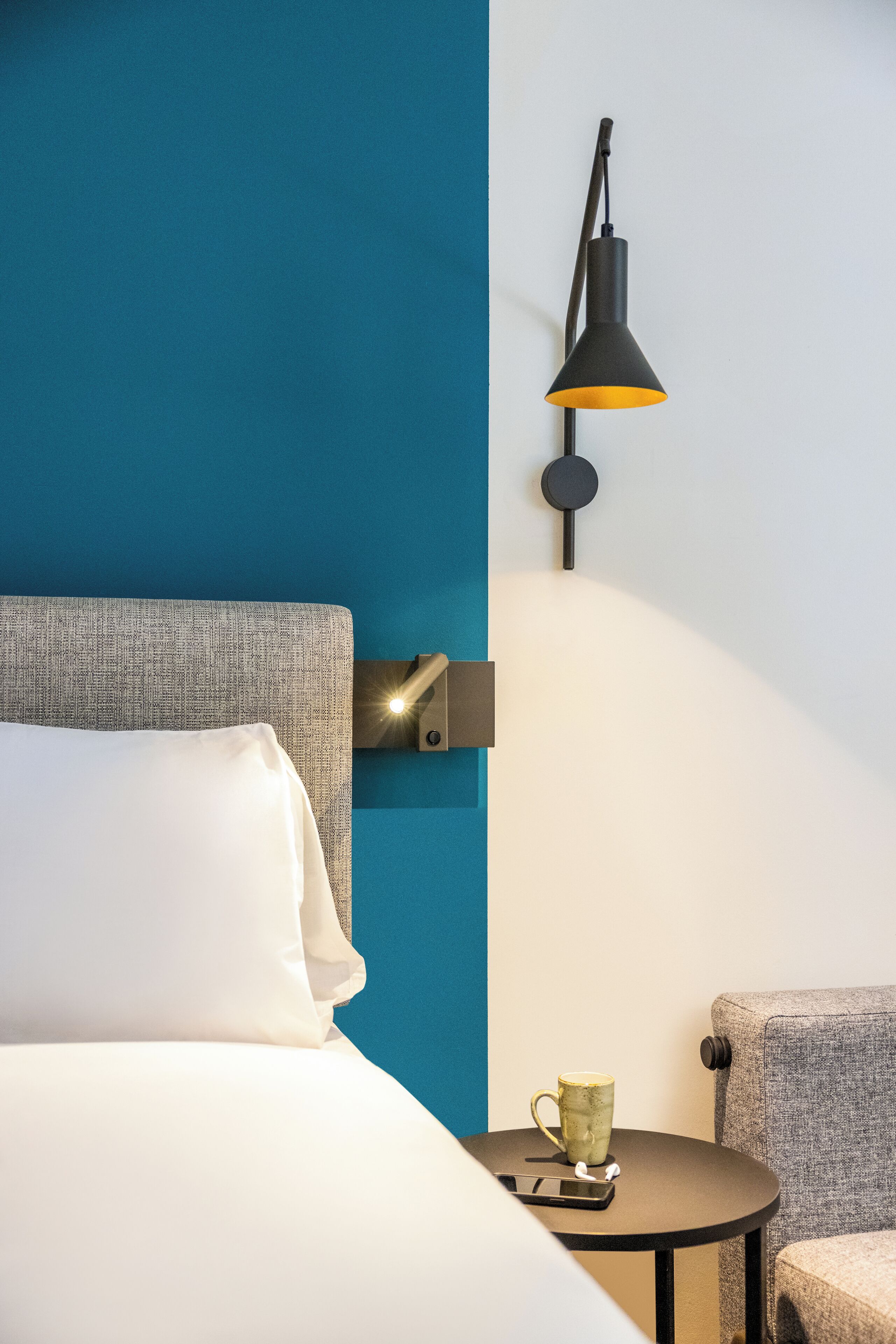 Una habitación de hotel con una cama, una mesita con una taza y un teléfono, una silla y una lámpara en la pared.