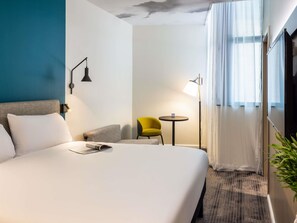 Une chambre d’hôtel avec un grand lit, une petite table ronde, une chaise jaune, un lampadaire et une vue sur l’extérieur par une fenêtre ornée de rideaux légers.