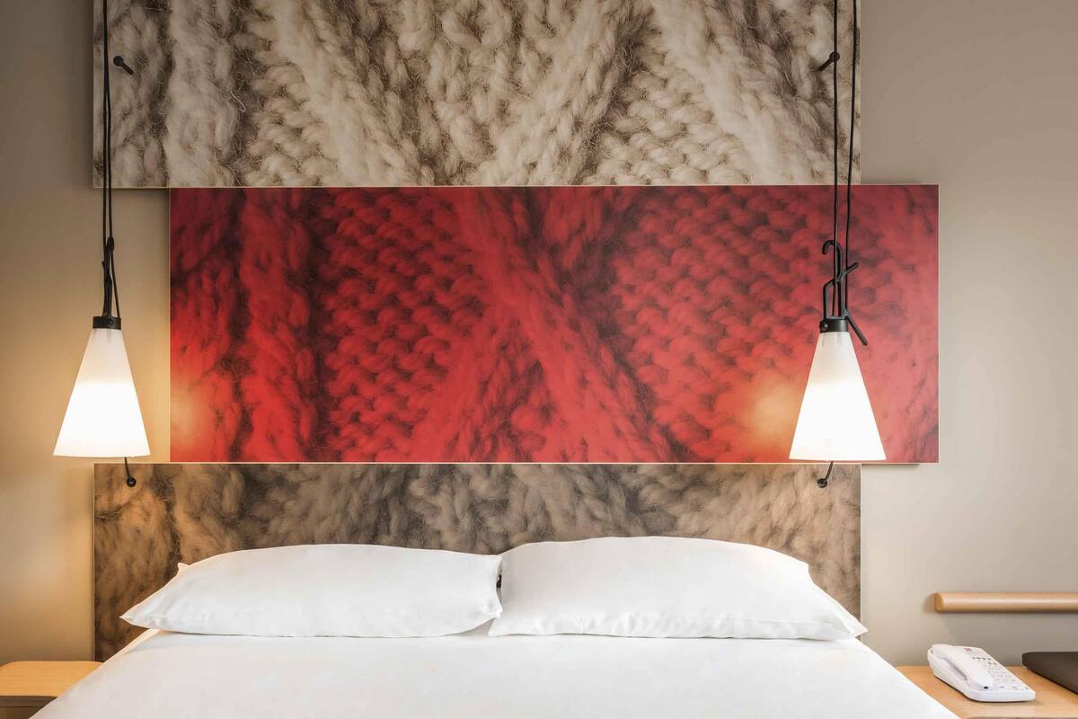 Ein Hotelzimmer mit einem Bett, zwei Pendelleuchten, einer Wandkunst mit rotem und beigem Muster sowie einem Nachttisch mit Telefon.