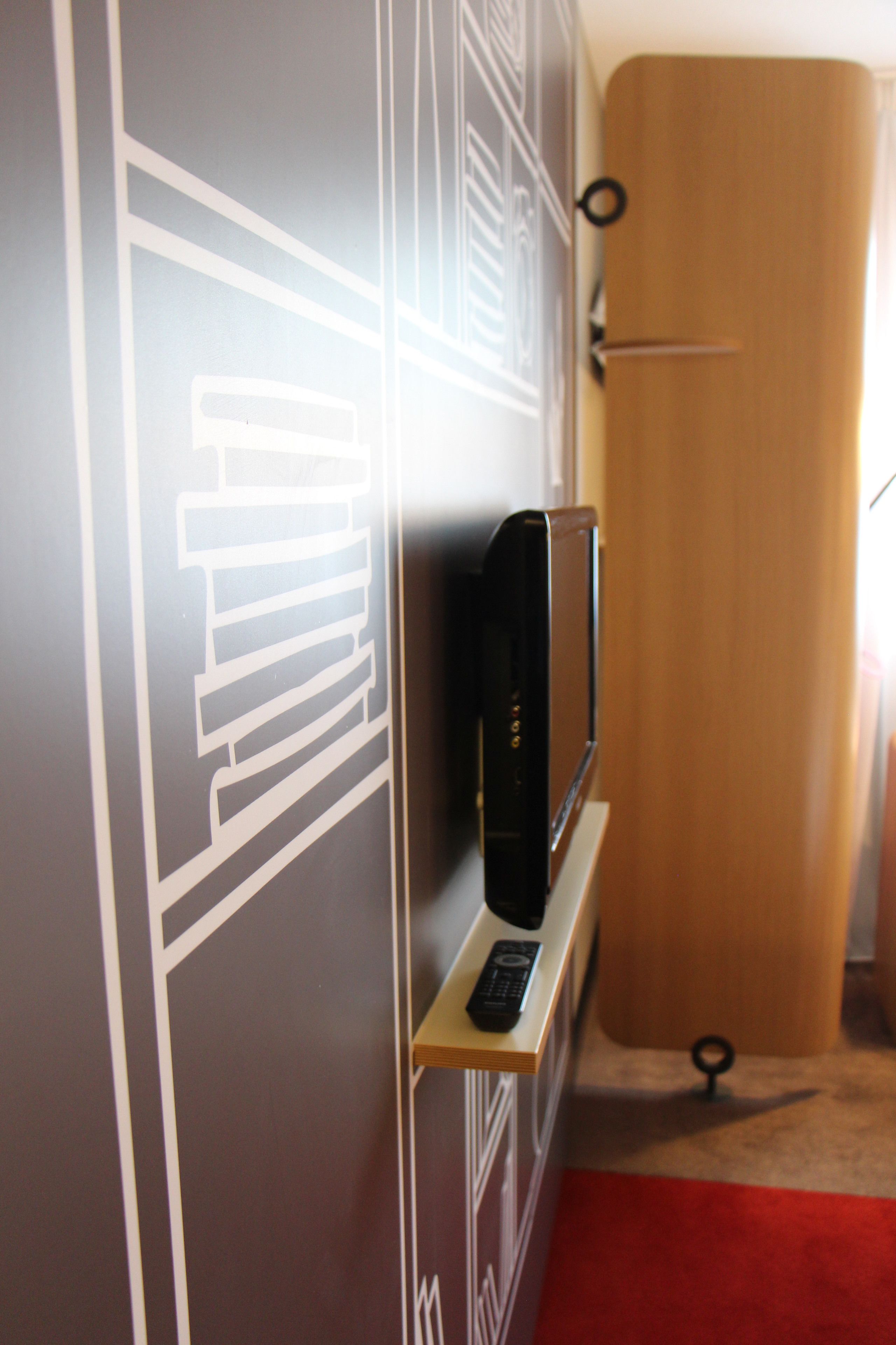 A hotel room with a television on a stand, a door with a handle, and a red carpet.