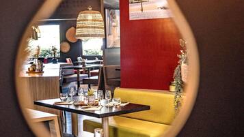 A restaurant interior with a mirrored wall reflecting the dining area, a yellow sofa, and a hanging chandelier.