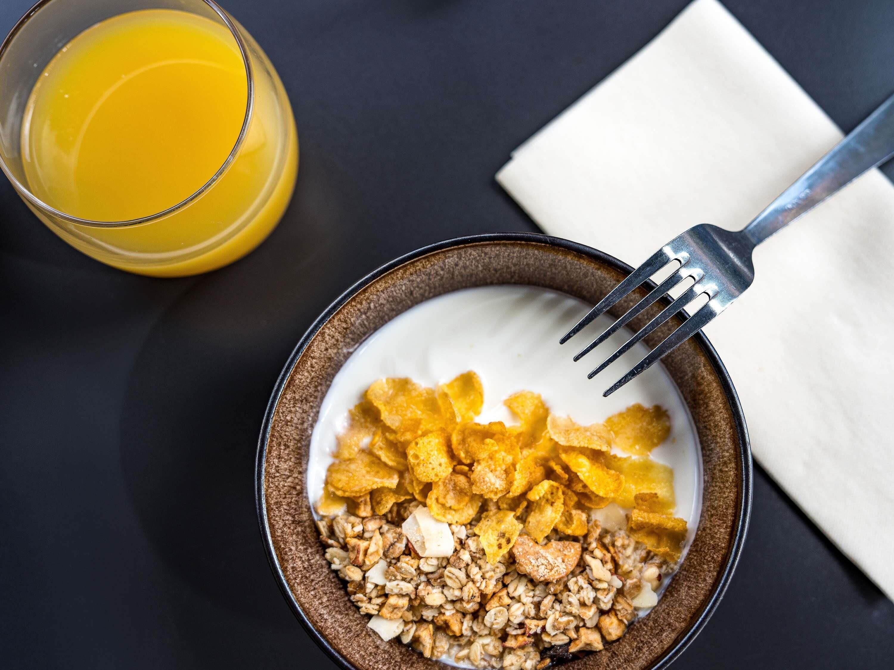 Eine Schüssel mit Frühstückscerealien, Obststücken und Granola, ein Glas Orangensaft und eine Gabel auf einer dunklen Oberfläche.
