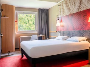 A hotel room with a bed, a desk, a chair, a wardrobe, a window with curtains, and a wall with a textured pattern. - ibis Arcachon La Teste (La Teste-de-Buch)