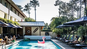 Una piscina con sillones de descanso y sombrillas, rodeada de árboles y un edificio con tejado de baldosas.
