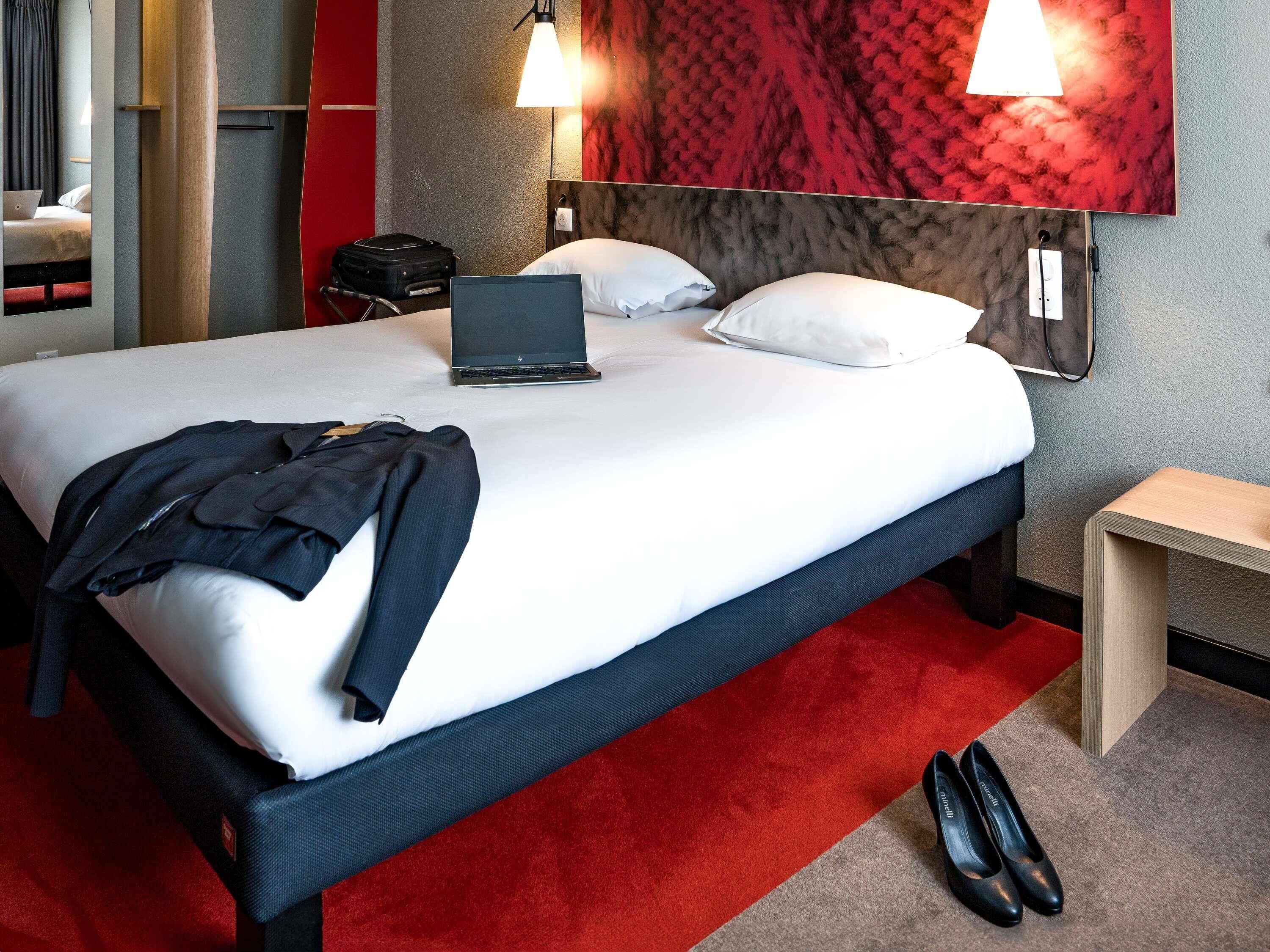 A neatly made bed with a laptop, a suitcase, and a pair of shoes on the floor.