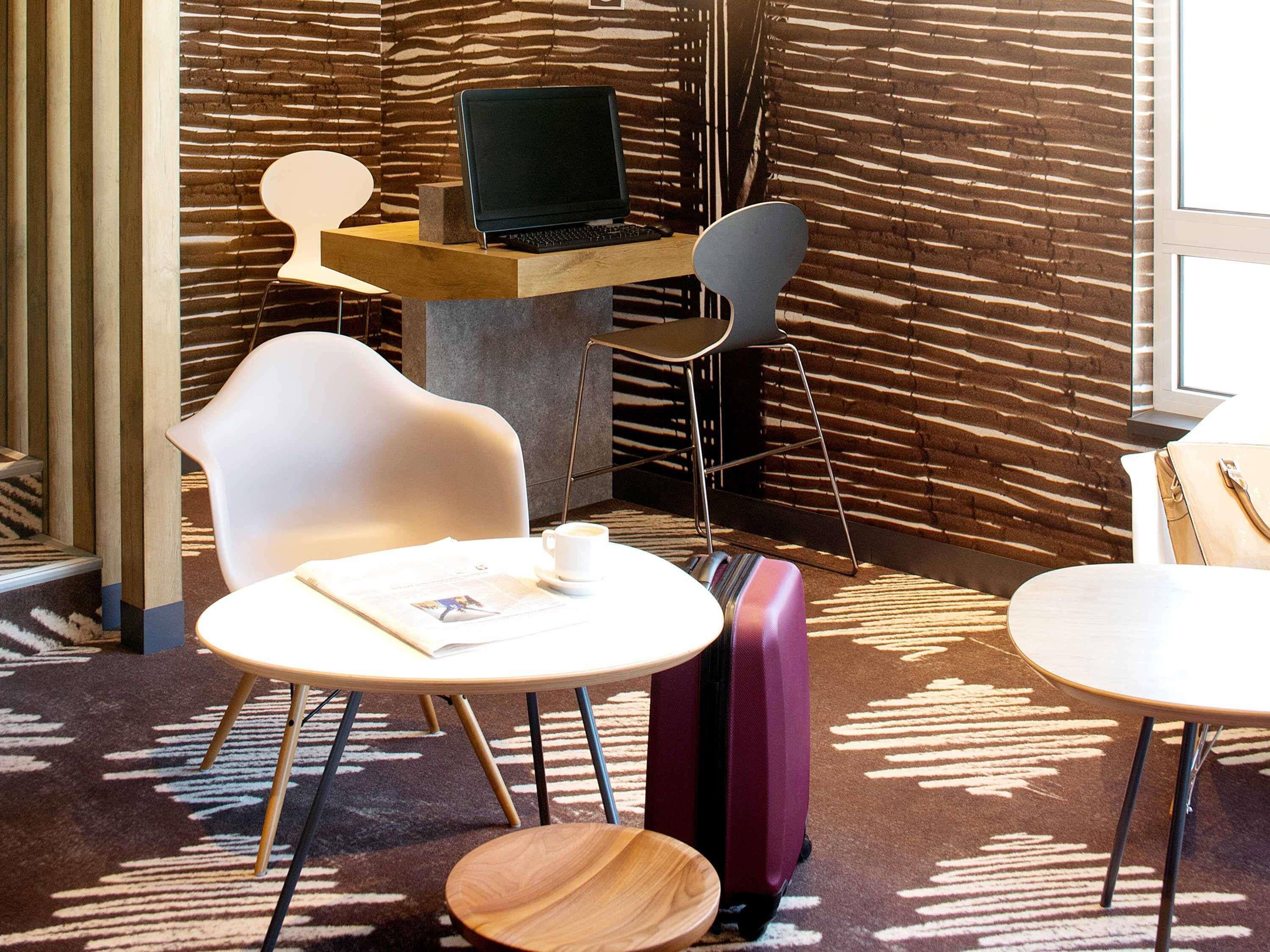 A modern hotel lobby with a wooden wall, a laptop on a desk, two white chairs, a purple suitcase, and a small wooden table.