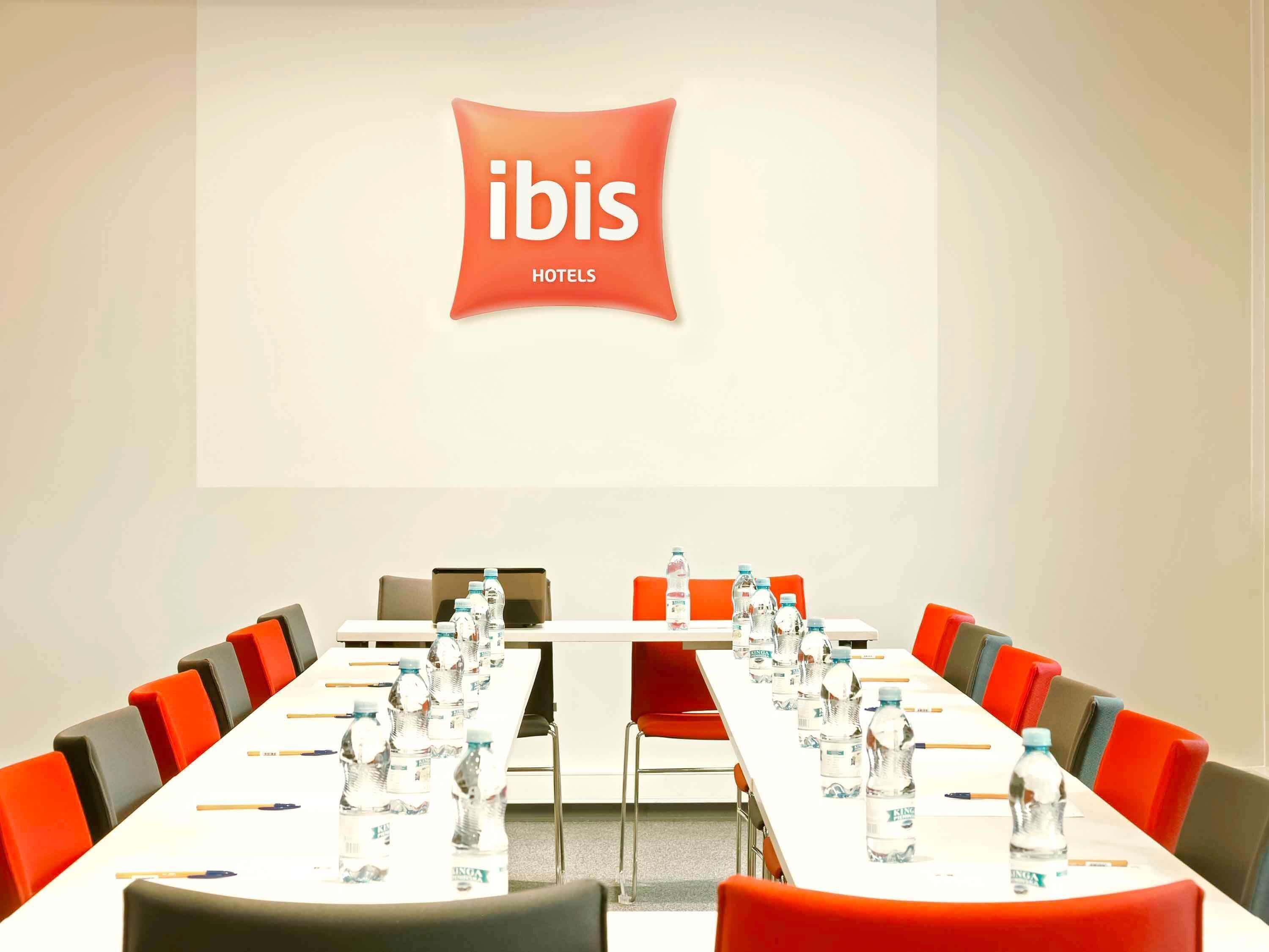 Un salón de conferencias con una larga mesa, sillas, botellas de agua y una pantalla mostrando el logotipo de ibis Hotels.