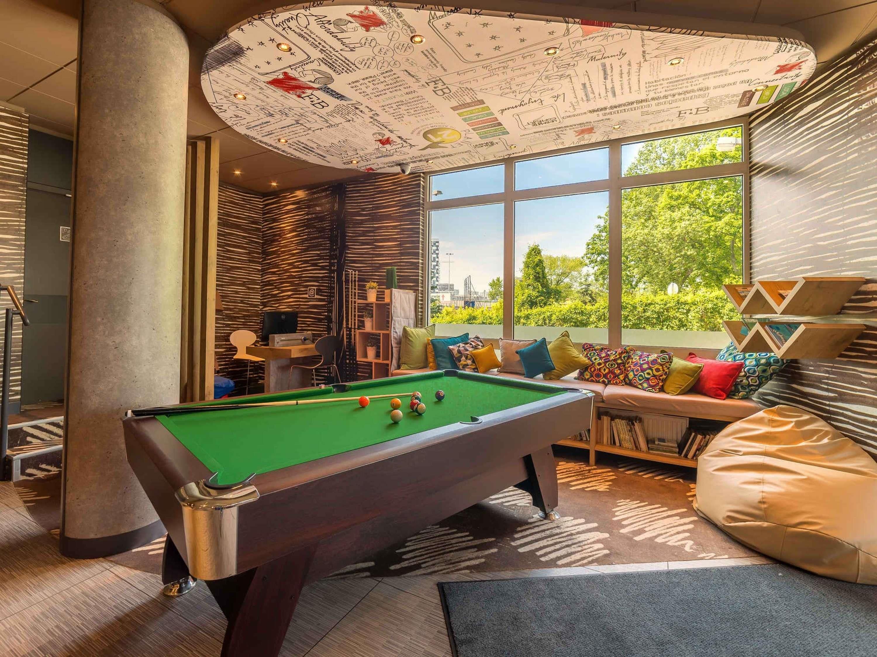 A pool table in a modern room with large windows, a bookshelf, and a unique ceiling design.