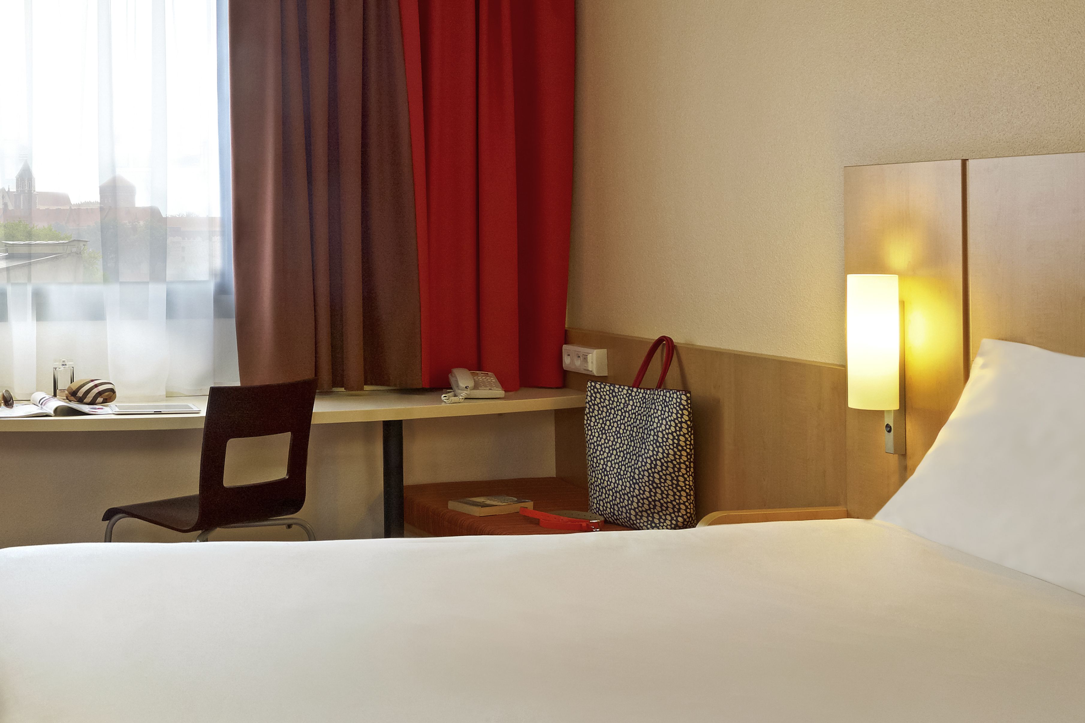 Quarto de hotel com cama, escrivaninha com cadeira, janela com cortinas, luminária e bolsa sobre a escrivaninha.