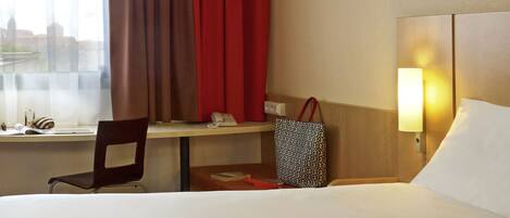 Quarto de hotel com cama, escrivaninha com cadeira, janela com cortinas, luminária e bolsa sobre a escrivaninha.