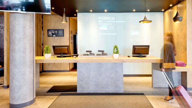 Un mostrador de recepción con dos monitores de ordenador, una planta y una pantalla digital que muestra información sobre las habitaciones.