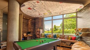 Una mesa de billar en una habitación moderna con ventanas amplias, una estantería y un diseño de techo original.