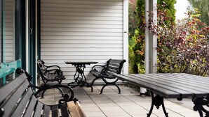 Une terrasse couverte avec deux tables et des bancs en métal noir, des murs recouverts de bardage blanc et un jardin agrémenté d’arbustes aux feuilles rouges.